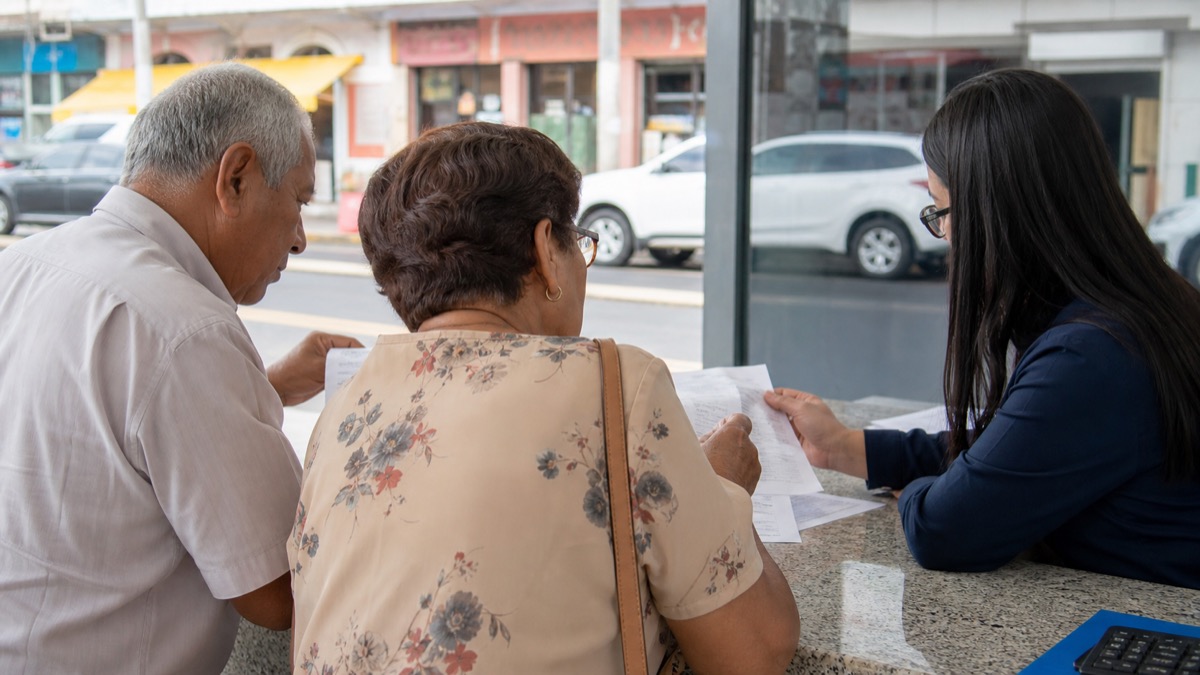older adults reviewing receipts with a consumer-service worker at a Panama storefront counter, no readable receipts