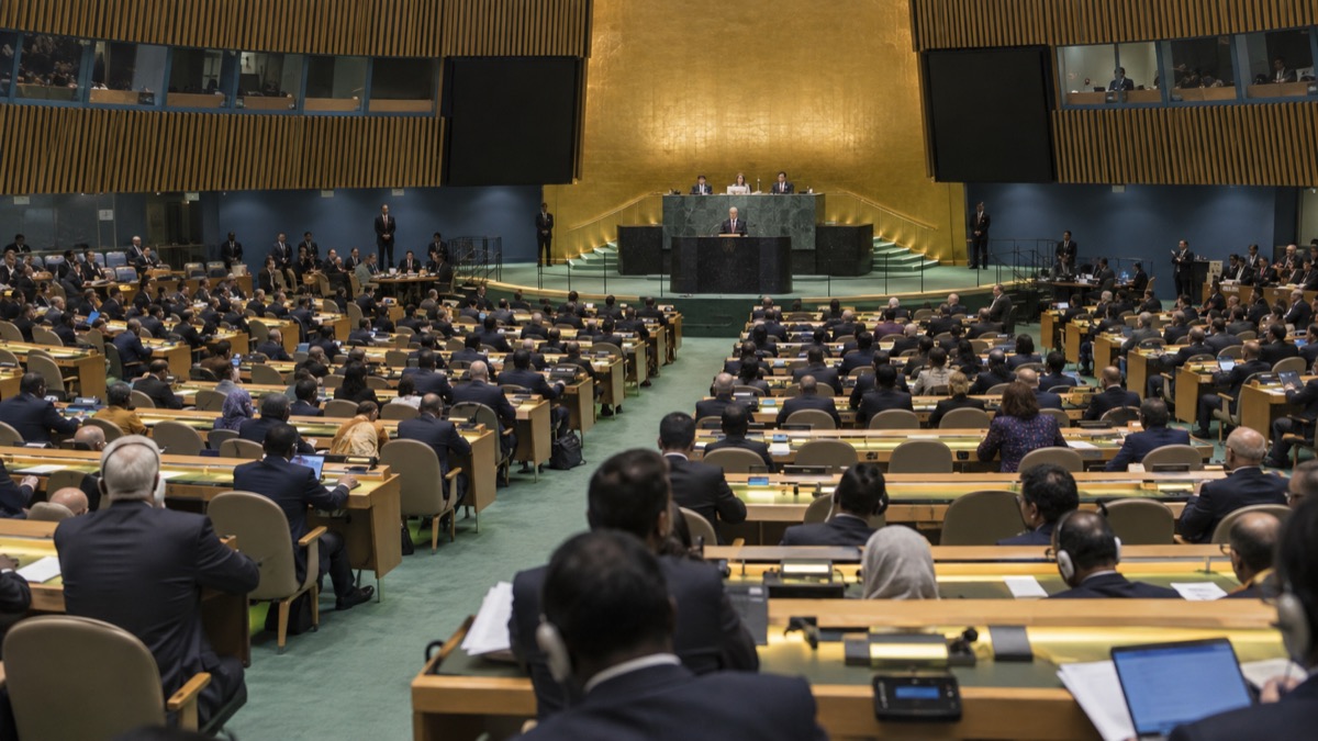 wide realistic editorial view of an international assembly hall with diverse delegates listening to a speaker at a distant podium, no flags, no readable text, no identifiable public figures